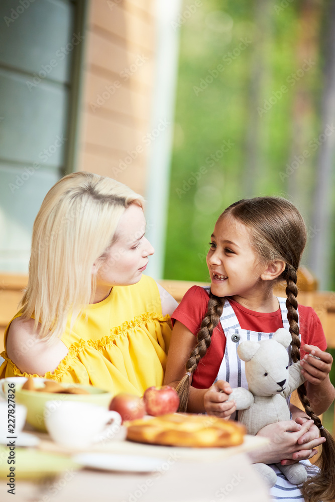 Cheerful little girl with teddy and her mother looking at each other during talk by breakfast in their country house