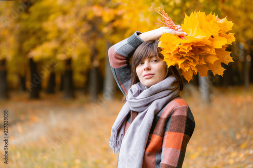 Portrait of young girl outdoor. Bouquet with yellow leafs in her hands. Brunette woman in autumn park with fashionable plaid coat and scarf.