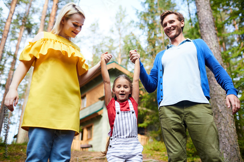 Cheerful little daughter holding by hands of her mom and dad during chill in the country