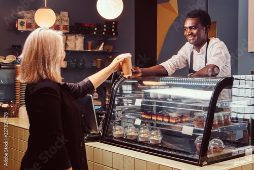 African American barista giving cup of coffee to his female client at the trendy coffee shop.