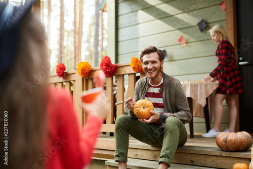 Young man with pumpkin sitting on terrace and talking to little daughter on background of his wife serving table for lunch photo