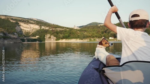 Slow motion a happy boy with her dog Jack Russell breed paddling on an inflatable kayak on the water of a large mountain lake on the background of a beautiful orange sunset. Family sports holiday