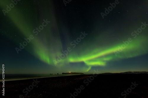 Bright green aurora borealis from Reynisfjara black sand beach in Iceland, looking towards the Dryholaey Peninsula and Lighthouse