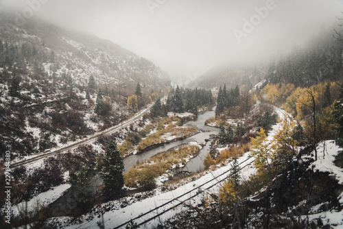 Landscape view of a railroad track and river surrounded by fall foliage during a snow storm. 