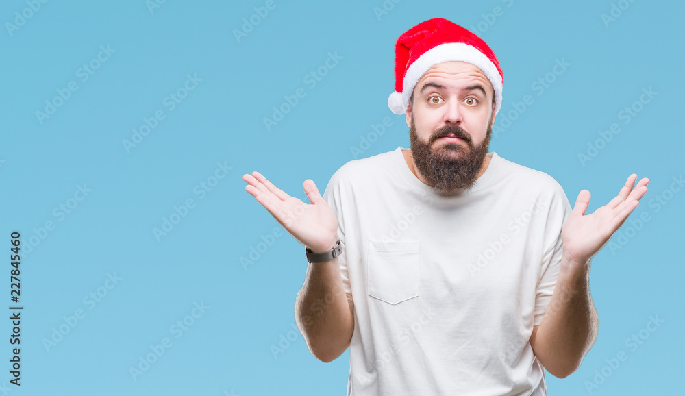 Young caucasian hipster man wearing christmas hat over isolated background clueless and confused expression with arms and hands raised. Doubt concept. Young caucasian hipster man wearing christmas hat over isolated background clueless and confused expression with arms and hands raised. Doubt concept.