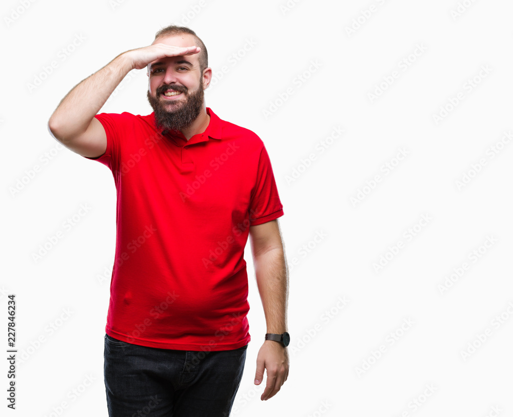 Young caucasian hipster man wearing red shirt over isolated background very happy and smiling looking far away with hand over head. Searching concept. Young caucasian hipster man wearing red shirt over isolated background very happy and smiling looking far away with hand over head. Searching concept.