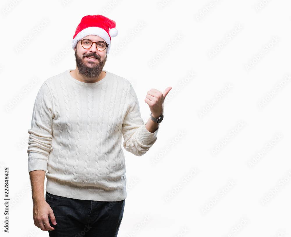 Naklejka premium Young caucasian man wearing christmas hat and glasses over isolated background smiling with happy face looking and pointing to the side with thumb up.