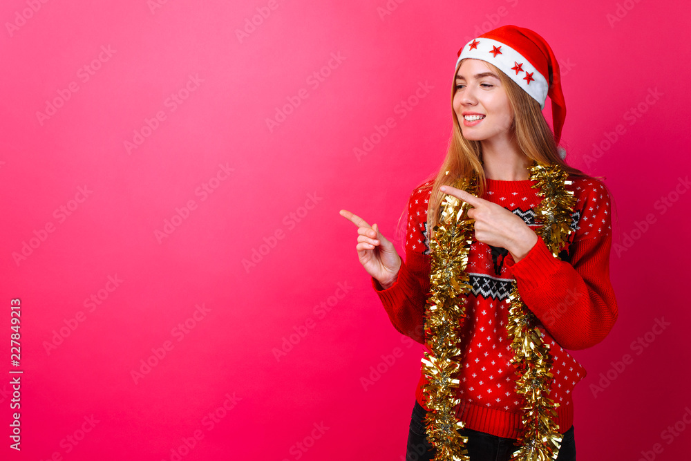 girl in a red sweater and Santa hat, with tinsel around her neck pointing at an empty space on a red background girl in a red sweater and Santa hat, with tinsel around her neck pointing at an empty space on a red background