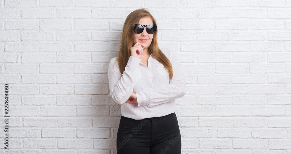 Young adult business woman standing over white brick wall wearing sunglasses serious face thinking about question, very confused idea Young adult business woman standing over white brick wall wearing sunglasses serious face thinking about question, very confused idea