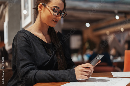 Young beautiful woman holding mobile phone in hands while waiting for her meal.