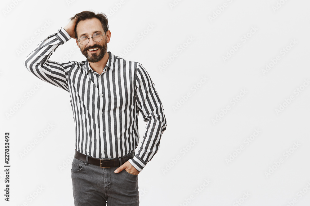 Man got carried away with stories from youth, apologizing with broad kind smile and scratching back of head while standing in formal outfit over gray wall, holding hand in pocket and gazing joyfully Man got carried away with stories from youth, apologizing with broad kind smile and scratching back of head while standing in formal outfit over gray wall, holding hand in pocket and gazing joyfully