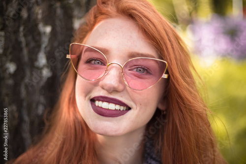 Redhead cheerful happy woman with freckles and dark lipstick wearing sunglasses outdoors in park sit under big tree on grass looking camera.
