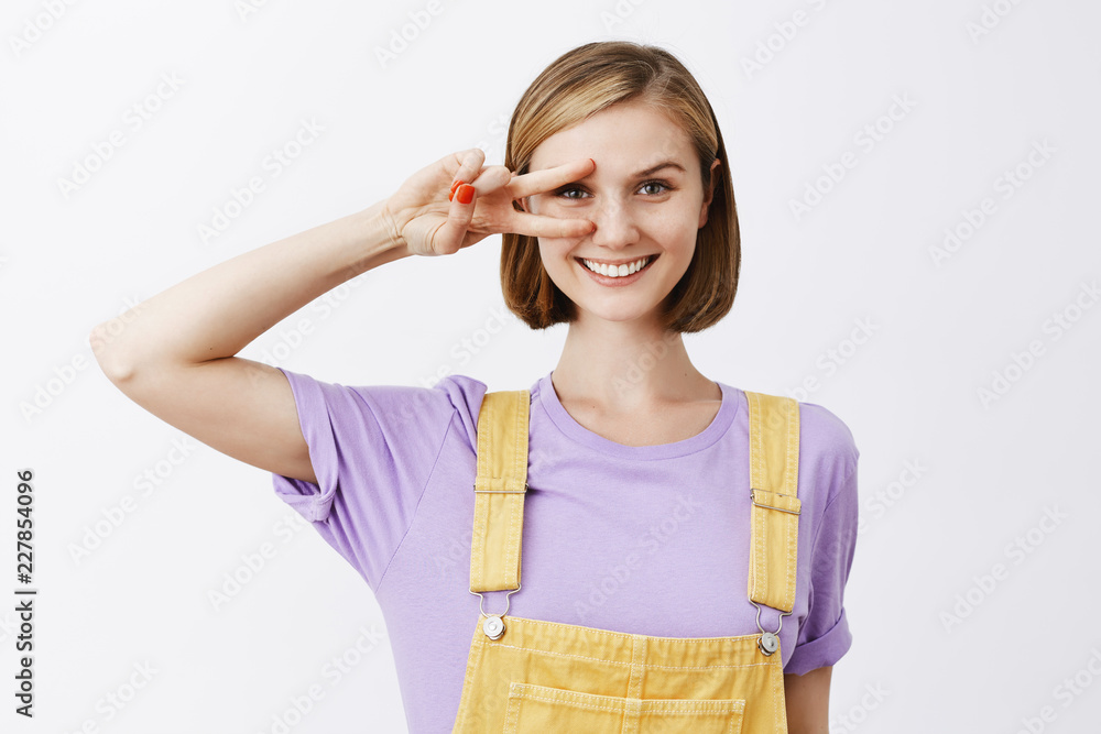 Bring peace not war. Portrait of charming bright and friendly-looking blond with short haircut, showing victory sign over eye and smiling joyfully, standing in cute overalls and gazing at camera Bring peace not war. Portrait of charming bright and friendly-looking blond with short haircut, showing victory sign over eye and smiling joyfully, standing in cute overalls and gazing at camera