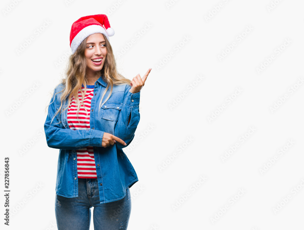 Beautiful young blonde woman wearing christmas hat over isolated background with a big smile on face, pointing with hand and finger to the side looking at the camera. Beautiful young blonde woman wearing christmas hat over isolated background with a big smile on face, pointing with hand and finger to the side looking at the camera.