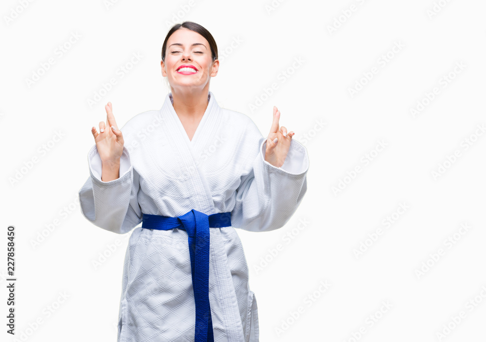Young beautiful woman wearing karate kimono uniform over isolated background smiling crossing fingers with hope and eyes closed. Luck and superstitious concept. Young beautiful woman wearing karate kimono uniform over isolated background smiling crossing fingers with hope and eyes closed. Luck and superstitious concept.