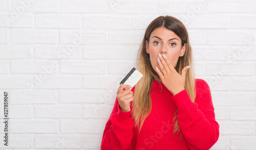 Young adult woman over white brick wall holding credit card cover mouth with hand shocked with shame for mistake, expression of fear, scared in silence, secret concept