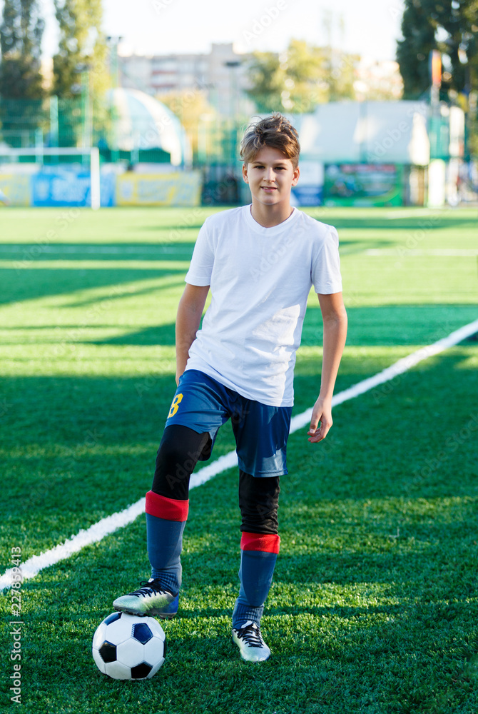 cute young boy in white blue sportswear stands next to classical black and white football ball on the stadium field. Soccer game, training, hobby concept.	
