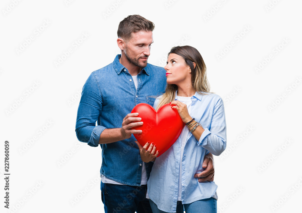 Young couple in love holding red heart over isolated background with a confident expression on smart face thinking serious
