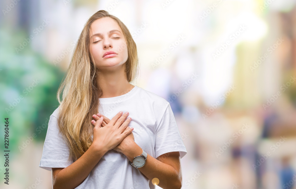Young beautiful blonde woman wearing casual white t-shirt over isolated background smiling with hands on chest with closed eyes and grateful gesture on face. Health concept.