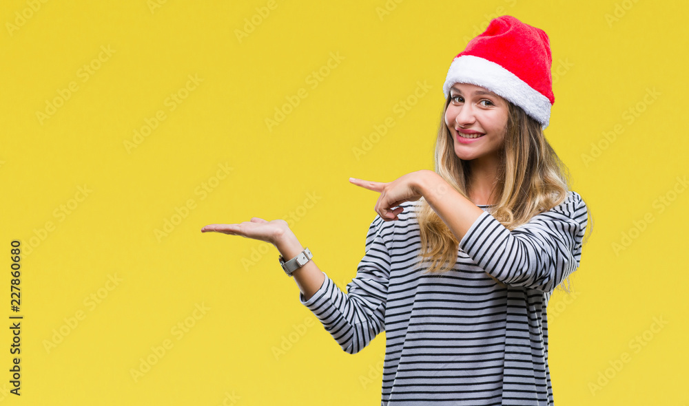 Young beautiful blonde woman wearing christmas hat over isolated background amazed and smiling to the camera while presenting with hand and pointing with finger. Young beautiful blonde woman wearing christmas hat over isolated background amazed and smiling to the camera while presenting with hand and pointing with finger.