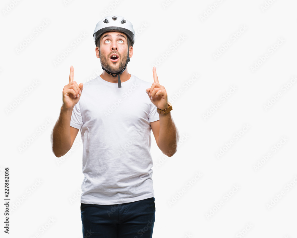 Young handsome man wearing cyclist safety helmet over isolated background amazed and surprised looking up and pointing with fingers and raised arms. Young handsome man wearing cyclist safety helmet over isolated background amazed and surprised looking up and pointing with fingers and raised arms.