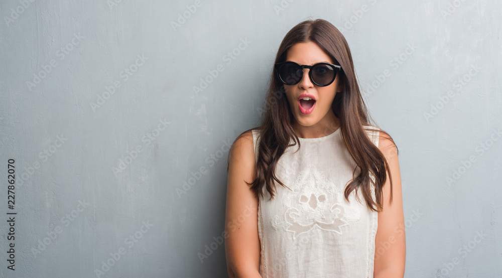 Young brunette woman over grunge grey wall wearing fashion sunglasses scared in shock with a surprise face, afraid and excited with fear expression Young brunette woman over grunge grey wall wearing fashion sunglasses scared in shock with a surprise face, afraid and excited with fear expression