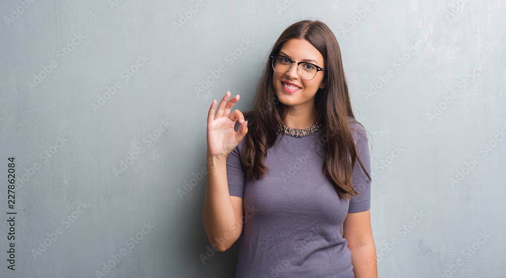 Young brunette woman over grunge grey wall wearing glasses doing ok sign with fingers, excellent symbol Young brunette woman over grunge grey wall wearing glasses doing ok sign with fingers, excellent symbol