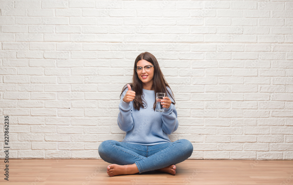 Young brunette woman sitting on the floor drinking glass of water happy with big smile doing ok sign, thumb up with fingers, excellent sign Young brunette woman sitting on the floor drinking glass of water happy with big smile doing ok sign, thumb up with fingers, excellent sign