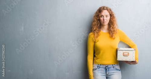 Young redhead woman over grey grunge wall holding box with a confident expression on smart face thinking serious