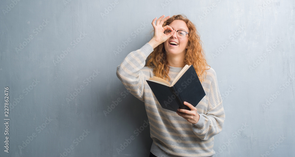 Young redhead woman over grey grunge wall reading a book with happy face smiling doing ok sign with hand on eye looking through fingers