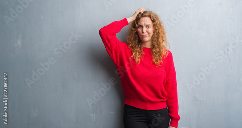 Young redhead woman over grey grunge wall wearing red sweater confuse and wonder about question. Uncertain with doubt  thinking with hand on head. Pensive concept.
