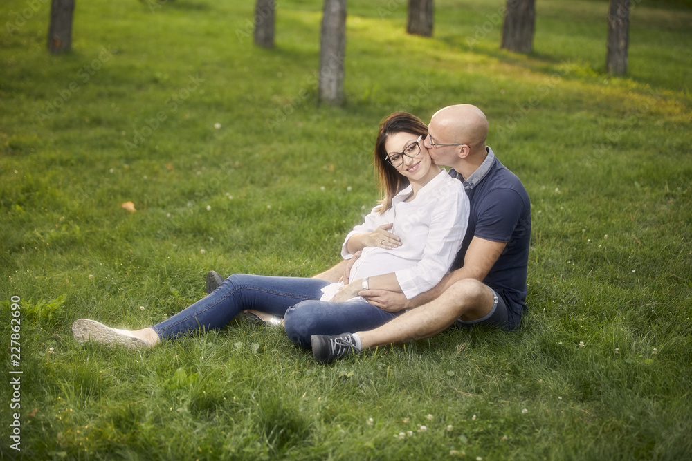 young couple sitting in grass field in park. smiling happy, man kissing pregnant woman. young couple sitting in grass field in park. smiling happy, man kissing pregnant woman.