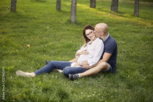 young couple sitting in grass field in park. smiling happy, man kissing pregnant woman.