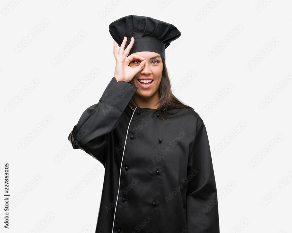Young hispanic cook woman wearing chef uniform with happy face smiling doing ok sign with hand on eye looking through fingers Young hispanic cook woman wearing chef uniform with happy face smiling doing ok sign with hand on eye looking through fingers