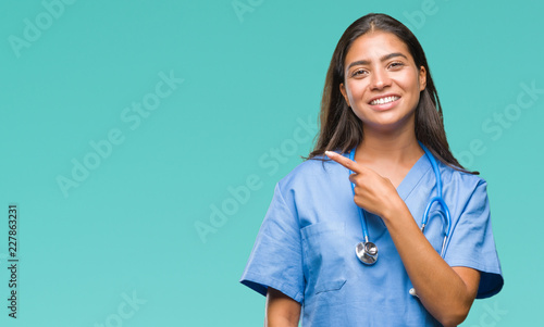 Young arab doctor surgeon woman over isolated background cheerful with a smile of face pointing with hand and finger up to the side with happy and natural expression on face looking at the camera. photo