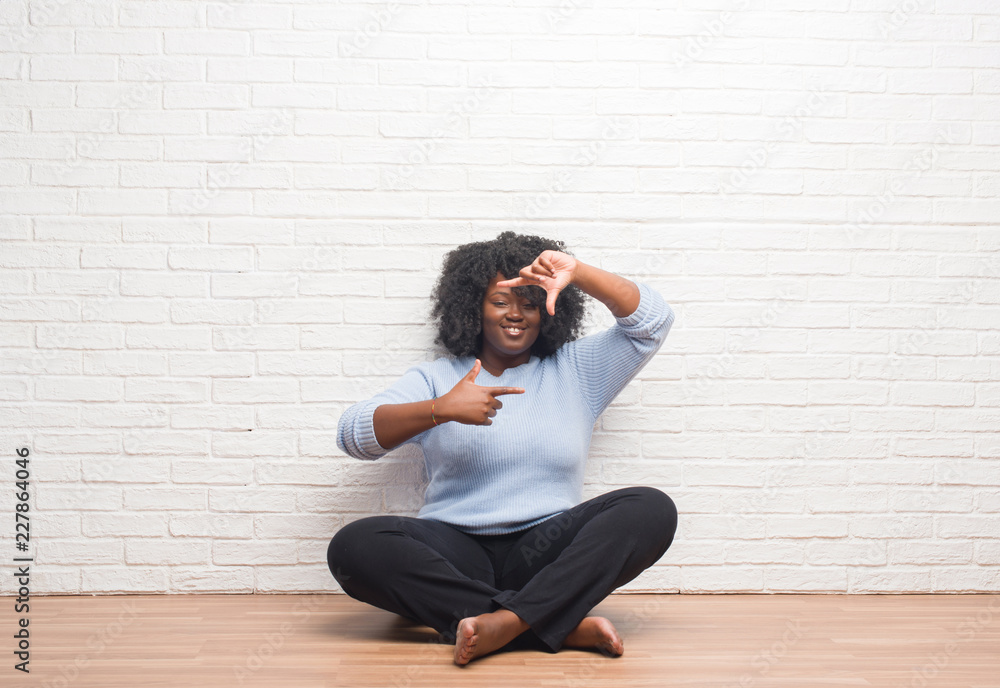Young african american woman sitting on the floor at home smiling making frame with hands and fingers with happy face. Creativity and photography concept. Young african american woman sitting on the floor at home smiling making frame with hands and fingers with happy face. Creativity and photography concept.