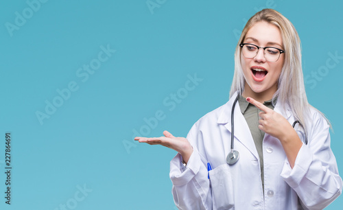 Young blonde doctor woman over isolated background amazed and smiling to the camera while presenting with hand and pointing with finger. photo