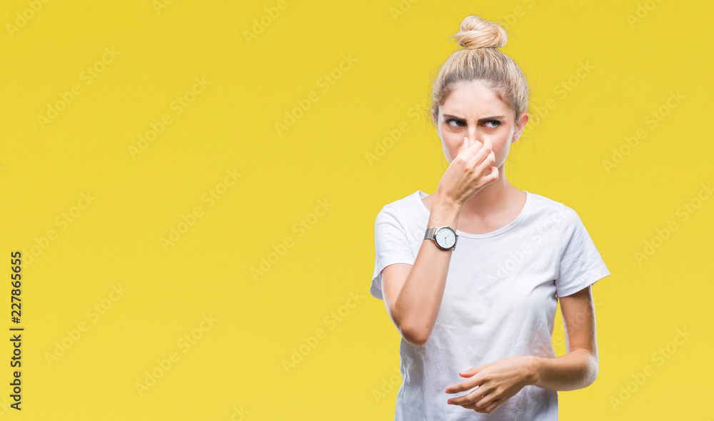 Young beautiful blonde woman wearing white t-shirt over isolated background smelling something stinky and disgusting, intolerable smell, holding breath with fingers on nose. Bad smells concept. Young beautiful blonde woman wearing white t-shirt over isolated background smelling something stinky and disgusting, intolerable smell, holding breath with fingers on nose. Bad smells concept.