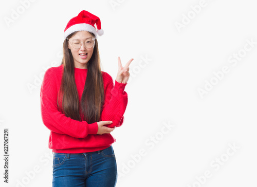 Young Chinese woman over isolated background wearing christmas hat smiling with happy face winking at the camera doing victory sign. Number two.