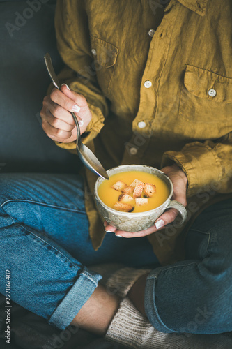 Female in yellow shirt and jeans sitting and eating Fall warming yellow pumpkin cream soup with croutons from ceramic mug. Autumn vegetarian, vegan, healthy comfort food eating concept