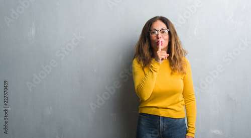 Middle age hispanic woman over grey wall wearing glasses asking to be quiet with finger on lips. Silence and secret concept.