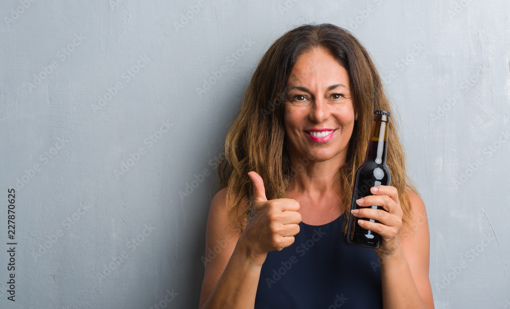 Middle age hispanic woman standing over grey grunge wall holding beer bottle happy with big smile doing ok sign, thumb up with fingers, excellent sign Middle age hispanic woman standing over grey grunge wall holding beer bottle happy with big smile doing ok sign, thumb up with fingers, excellent sign