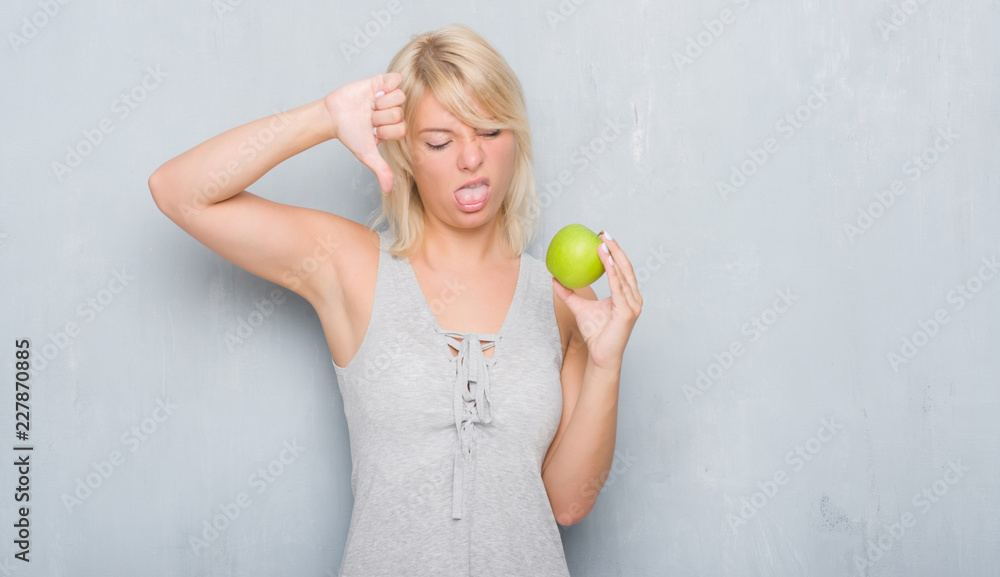 Adult caucasian woman over grunge grey wall eating green apple with angry face, negative sign showing dislike with thumbs down, rejection concept Adult caucasian woman over grunge grey wall eating green apple with angry face, negative sign showing dislike with thumbs down, rejection concept