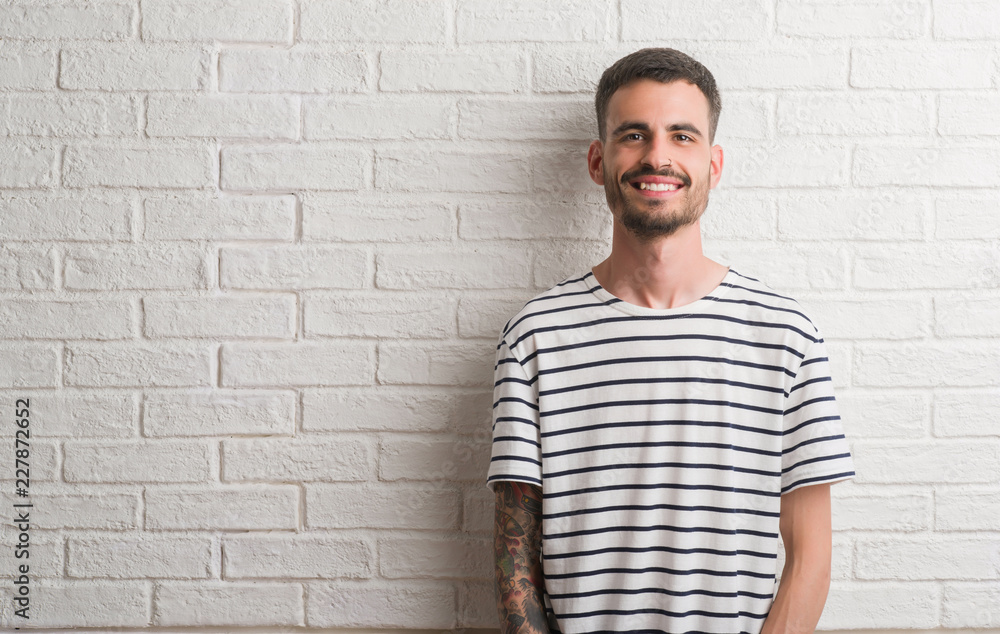 Young adult man standing over white brick wall with a happy face standing and smiling with a confident smile showing teeth Young adult man standing over white brick wall with a happy face standing and smiling with a confident smile showing teeth