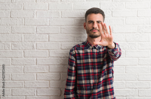 Young adult man standing over white brick wall doing stop sing with palm of the hand. Warning expression with negative and serious gesture on the face.