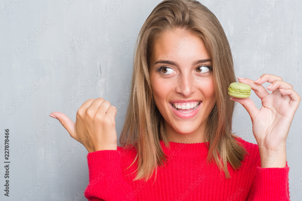 Beautiful young woman over grunge grey wall eating green macaron pointing and showing with thumb up to the side with happy face smiling Beautiful young woman over grunge grey wall eating green macaron pointing and showing with thumb up to the side with happy face smiling