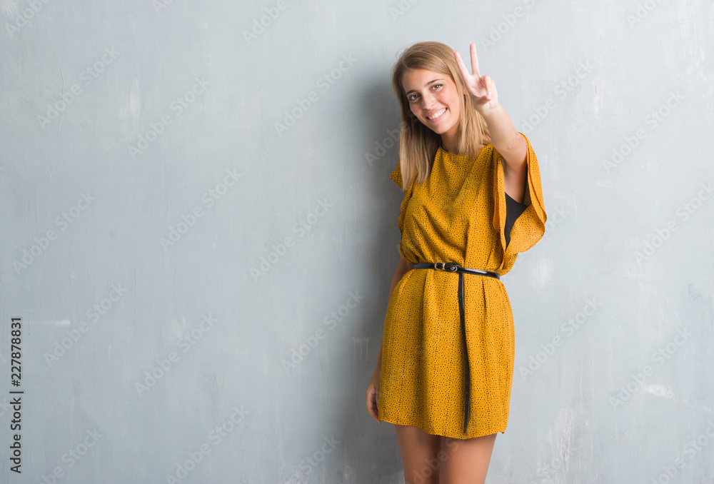 Beautiful young woman standing over grunge grey wall wearing a dress smiling looking to the camera showing fingers doing victory sign. Number two. Beautiful young woman standing over grunge grey wall wearing a dress smiling looking to the camera showing fingers doing victory sign. Number two.