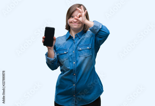 Young adult woman with down syndrome showing smartphone screen over isolated background with happy face smiling doing ok sign with hand on eye looking through fingers