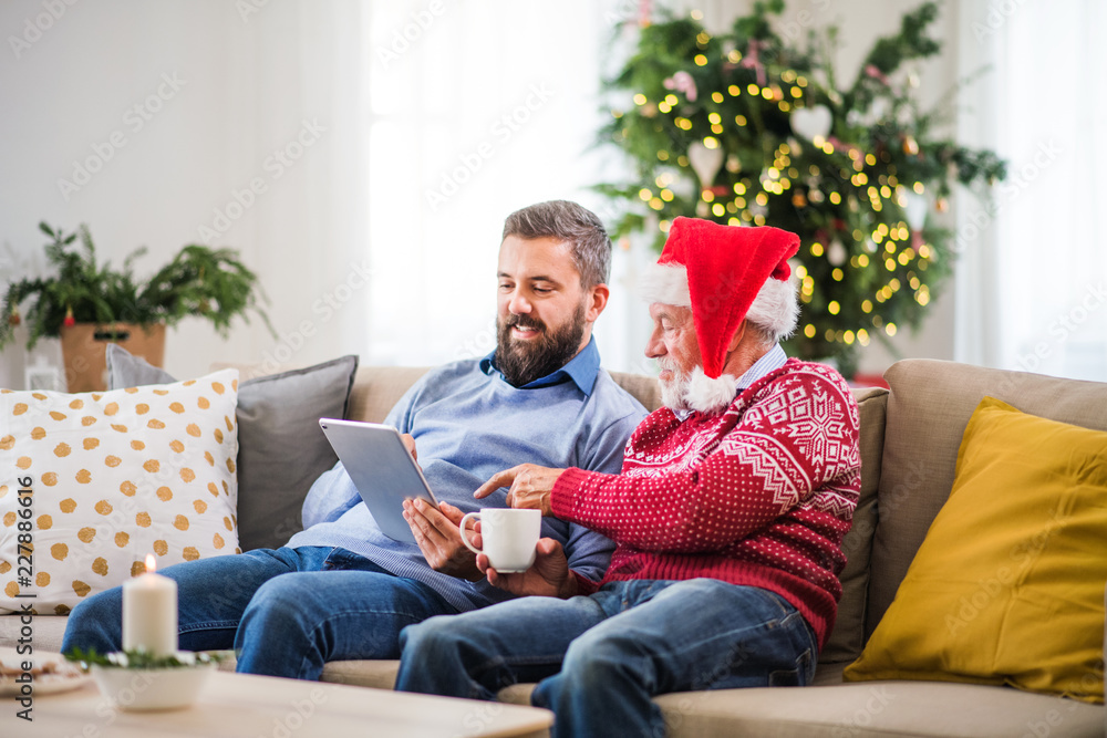 A senior father and adult son with tablet sitting on a sofa at home at Christmas time.