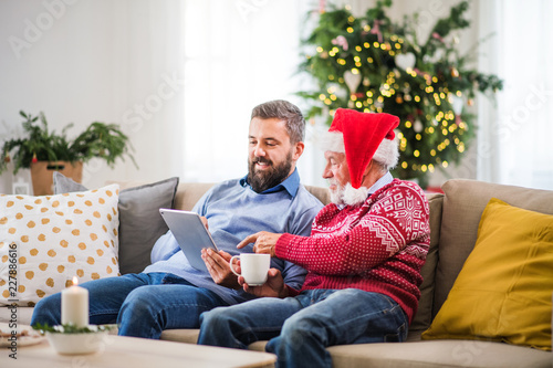 A senior father and adult son with tablet sitting on a sofa at home at Christmas time. © Halfpoint
